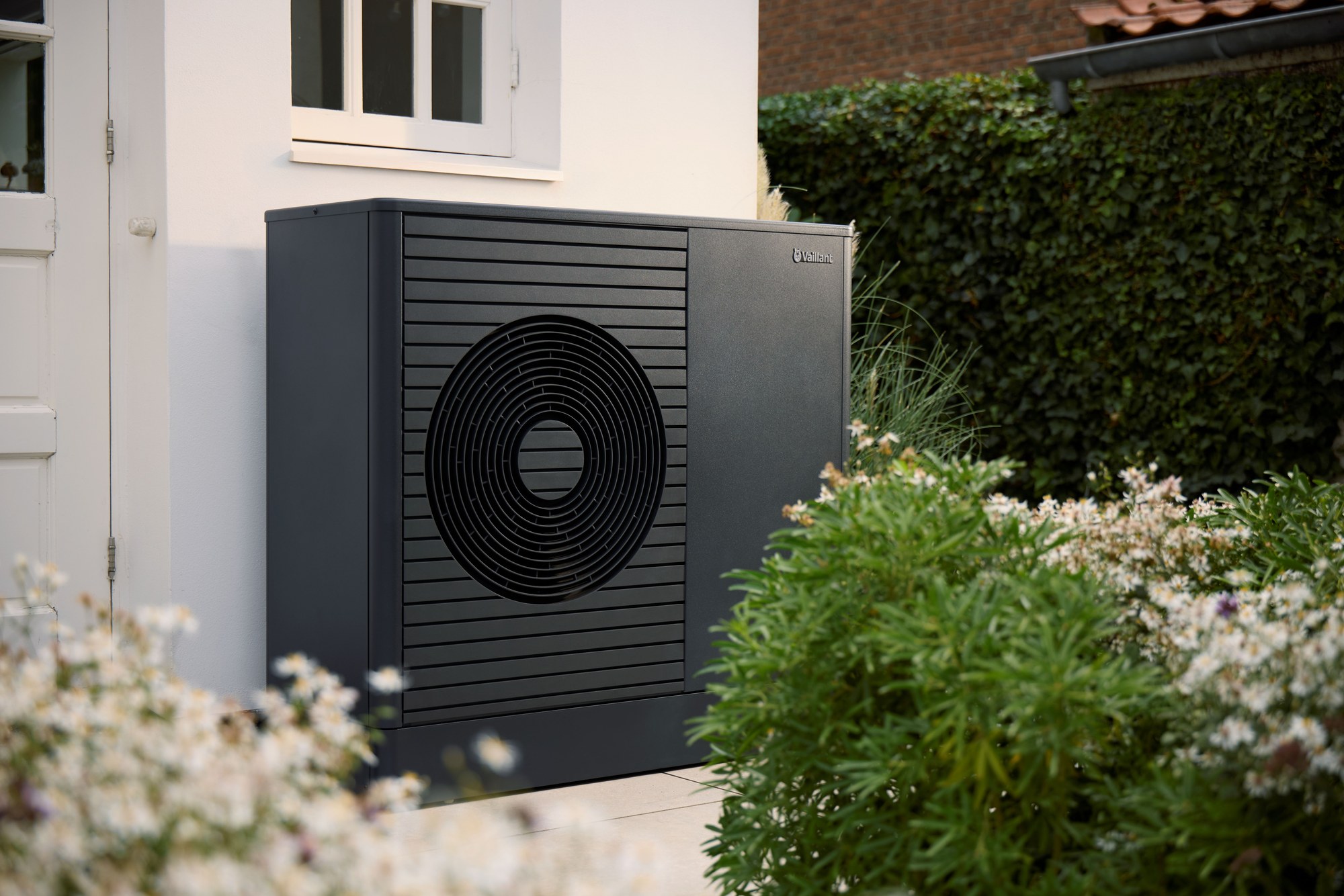 Sort Vaillant aroTHERM plus varmepumpe placeret ved en hvid husmur blandt blomster og grøn beplantning.